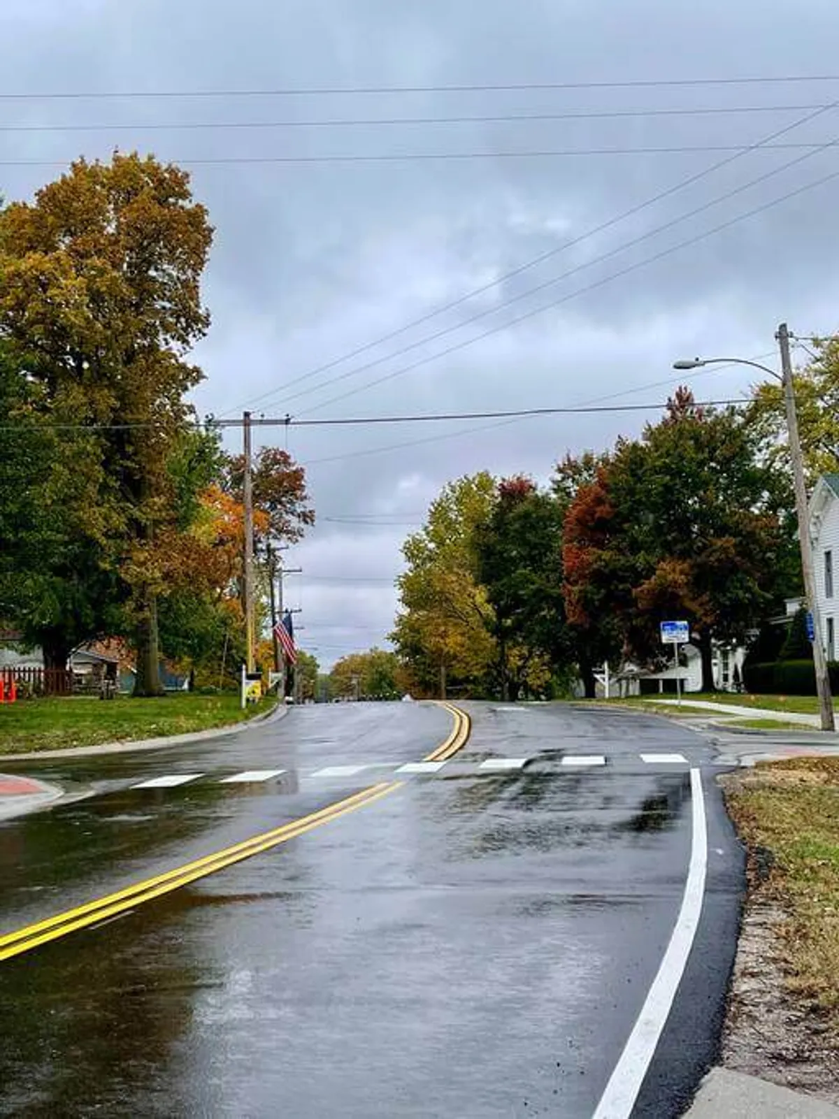 This image depicts a quiet neighborhood street with wet pavement, autumn-colored trees lining the road, an American flag, and visible street signs. The serene atmosphere hints at walkability and year-round outdoor enjoyment, with a touch of small-town charm.