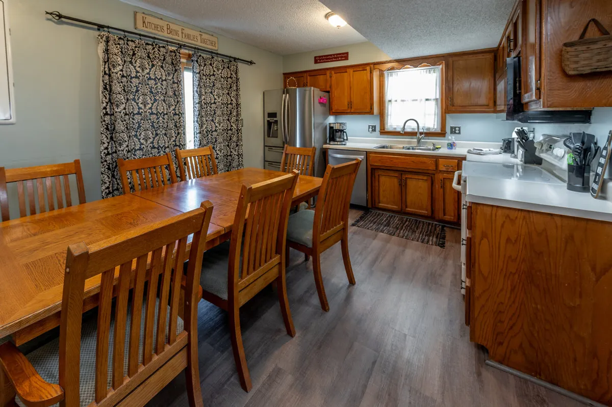 A bright kitchen and dining area with a neatly set table, inviting kitchen cabinets, and sunlit curtains framing a window. The setup suggests group gatherings, shared meals, and a natural connection to the outside—possibly through the window or an adjacent door.