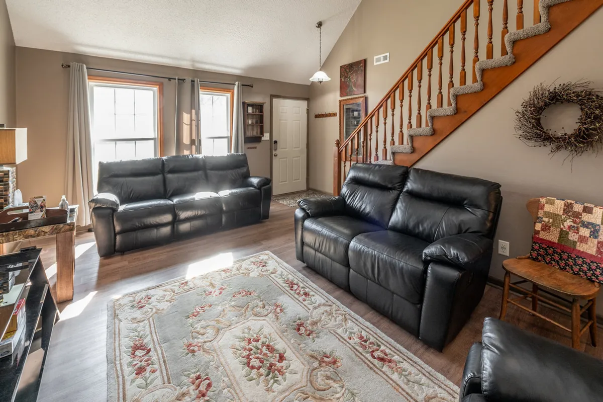 Another perspective on the spacious living zone, this image highlights plush black sofas, a vibrant area rug, and the staircase, all bathed in natural light. The space feels open and inviting, ideal for quiet afternoons or lively gatherings.