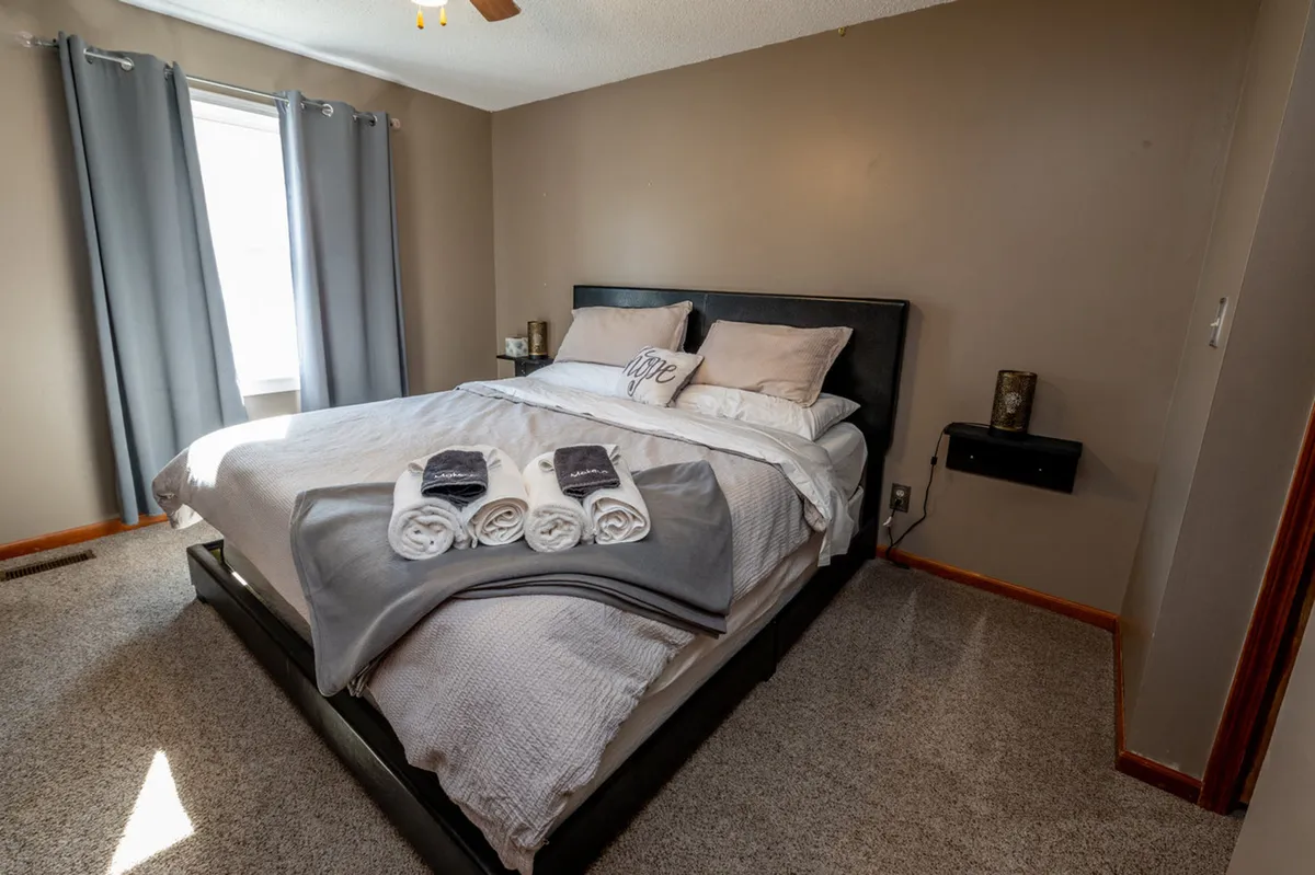 An inviting bedroom with a neatly made bed, decorative pillows, warm blankets, and a large window with curtains. The nightstand is adorned with tasteful decor, and sunlight streams into the space, evoking a cozy and restful vibe.