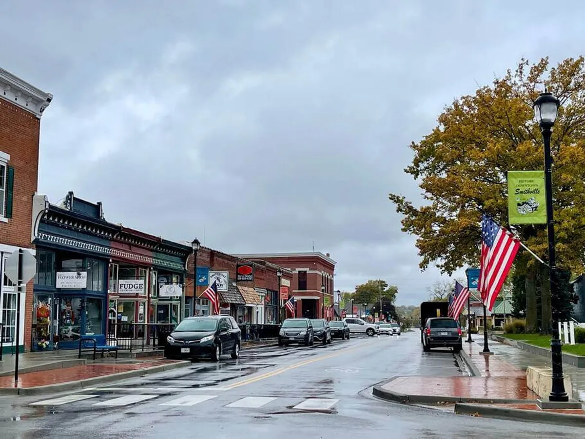 A lively streetscape with historic storefronts adorned with American flags, clean sidewalks, and classic lamp posts. The scene conveys a sense of community and offers hints of local shops, cafes, and event spaces.