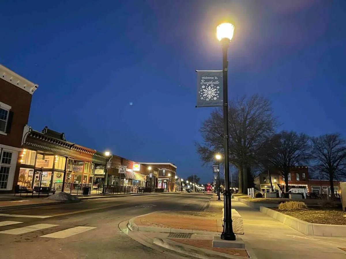 A charming downtown street scene at dusk, featuring warmly lit shopfronts, a welcoming banner stretched across the road, and a classic street lamp. The area appears lively but safe, with hints of local businesses that invite exploration and family-friendly outings.