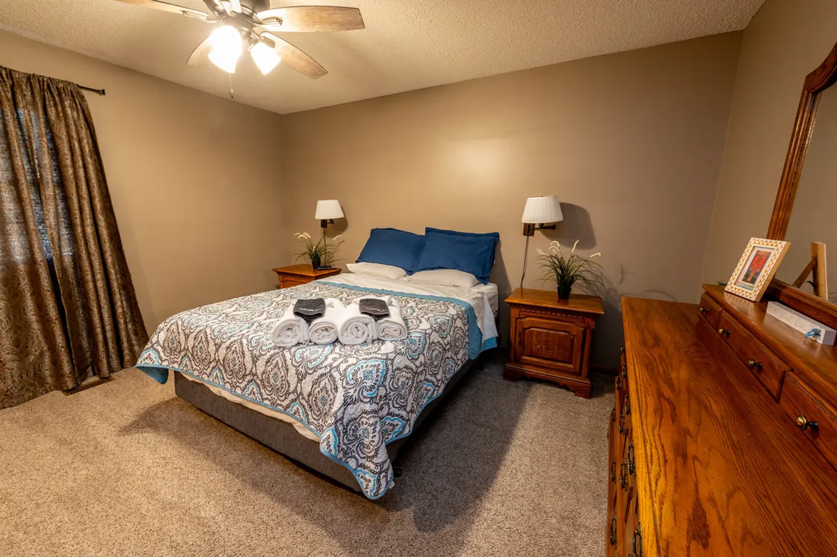 A tranquil bedroom featuring a bed with a patterned bedspread, flanked by nightstands with lamps. A wooden dresser sits against the wall, and soft natural light filters through curtains, creating a peaceful atmosphere ideal for relaxation.